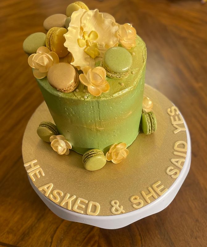 Sage green engagement cake topped with macarons and gold flowers, lettered He asked and she said yes
